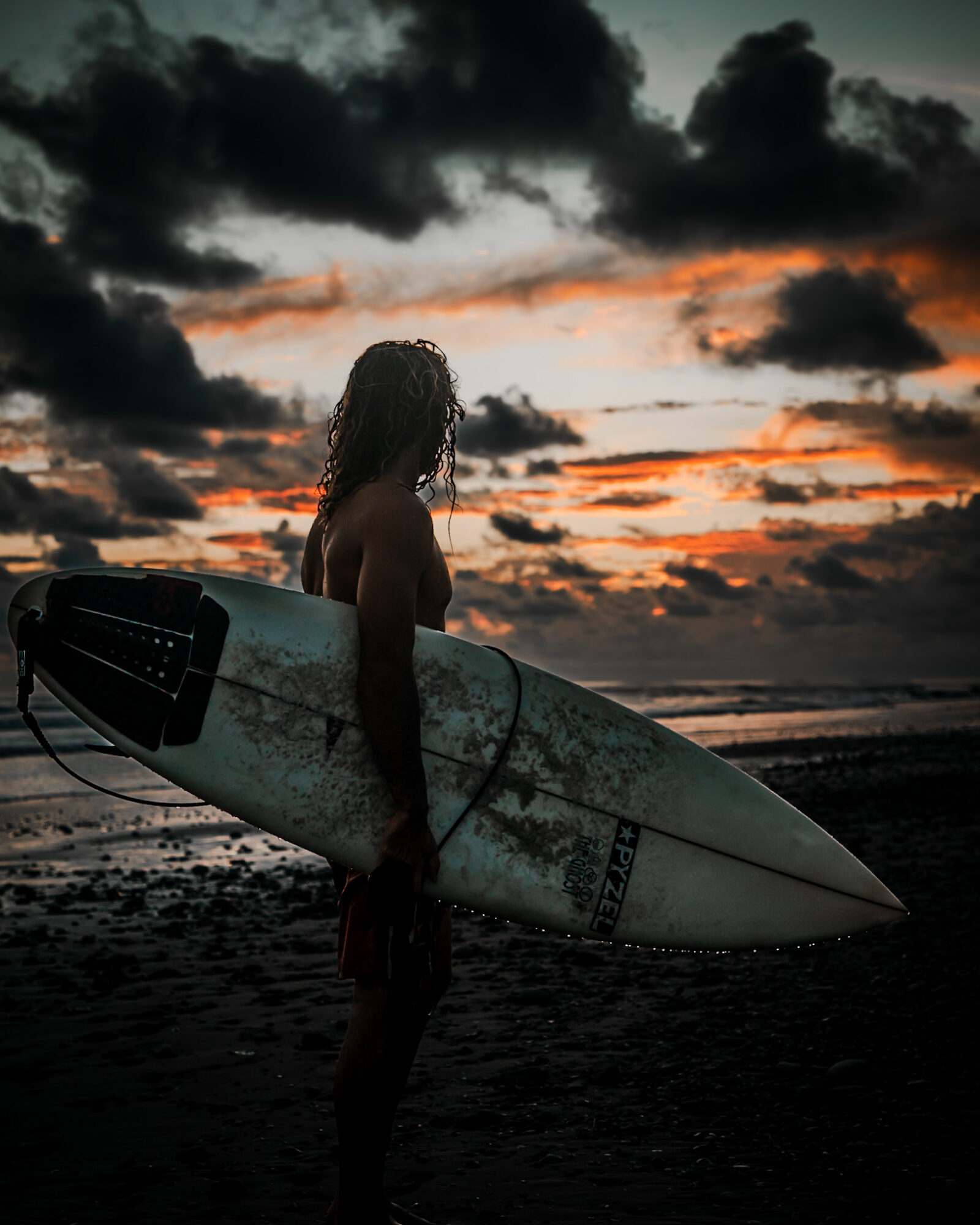 Person holding surfboard on beach during sunset with dark clouds in sky.