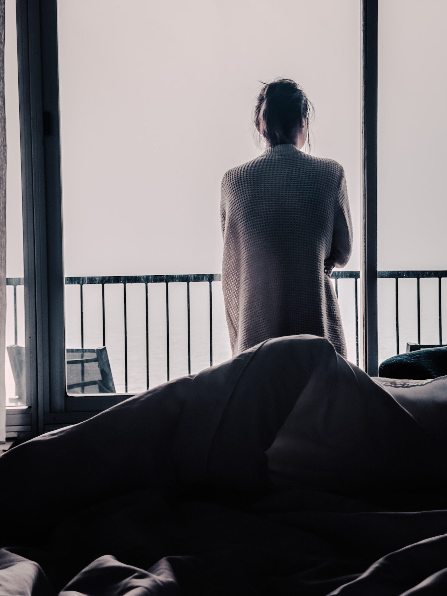 Person standing on balcony, looking outside, with railing and bright sky in background.