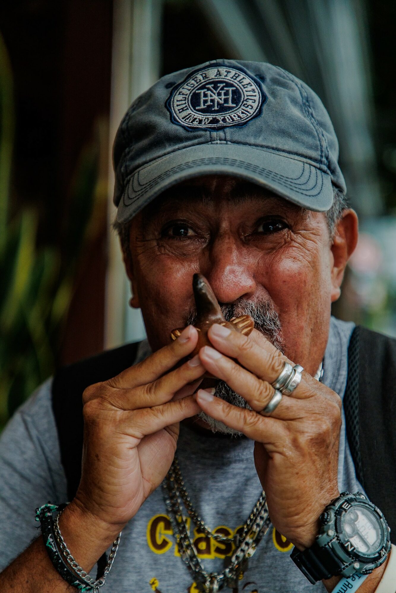 Elderly person wearing a blue cap, holding a small object near their mouth, with rings and a watch visible.