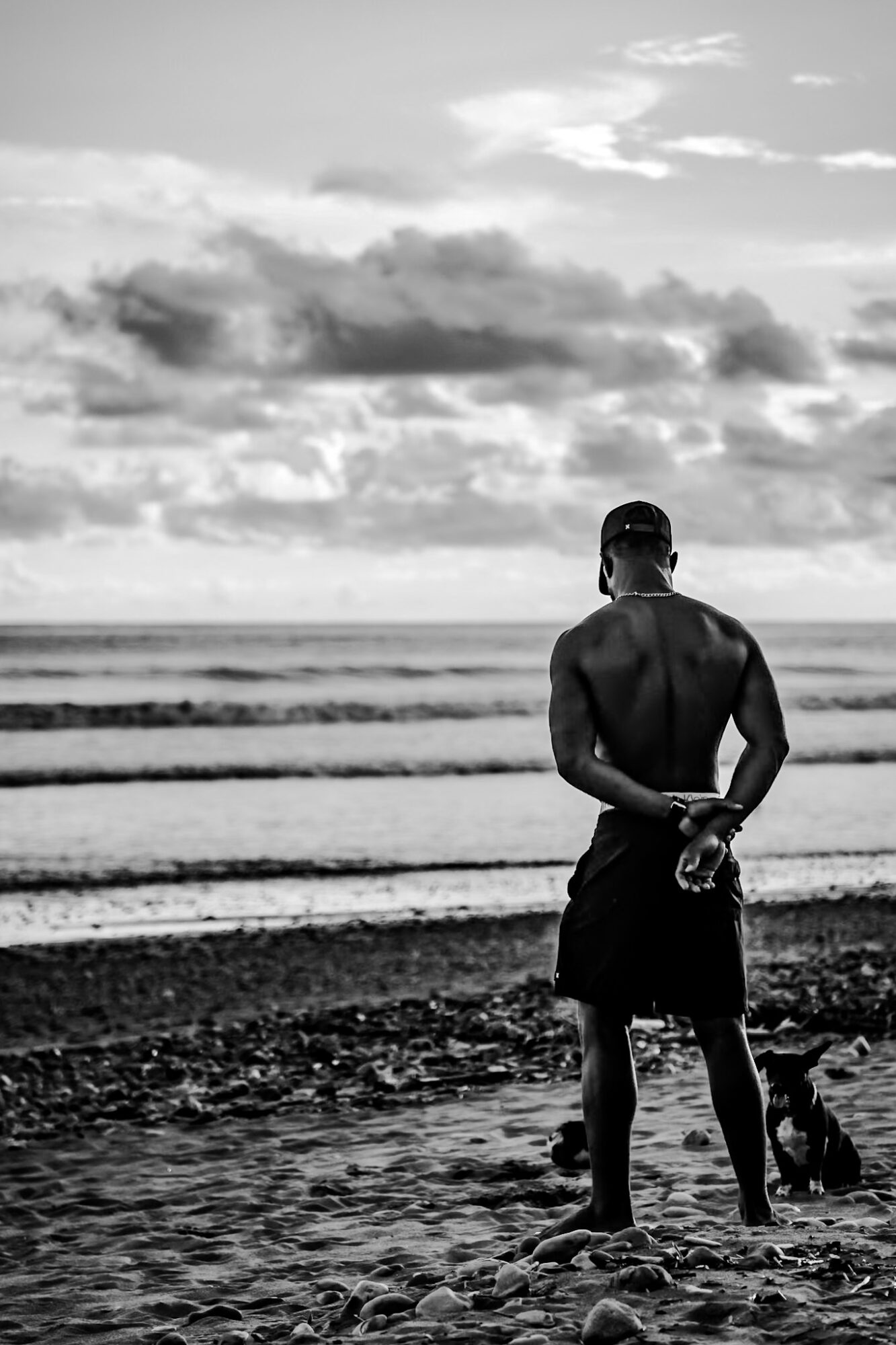 Man standing on beach facing ocean, holding shoes behind back, cloudy sky above, waves in background.