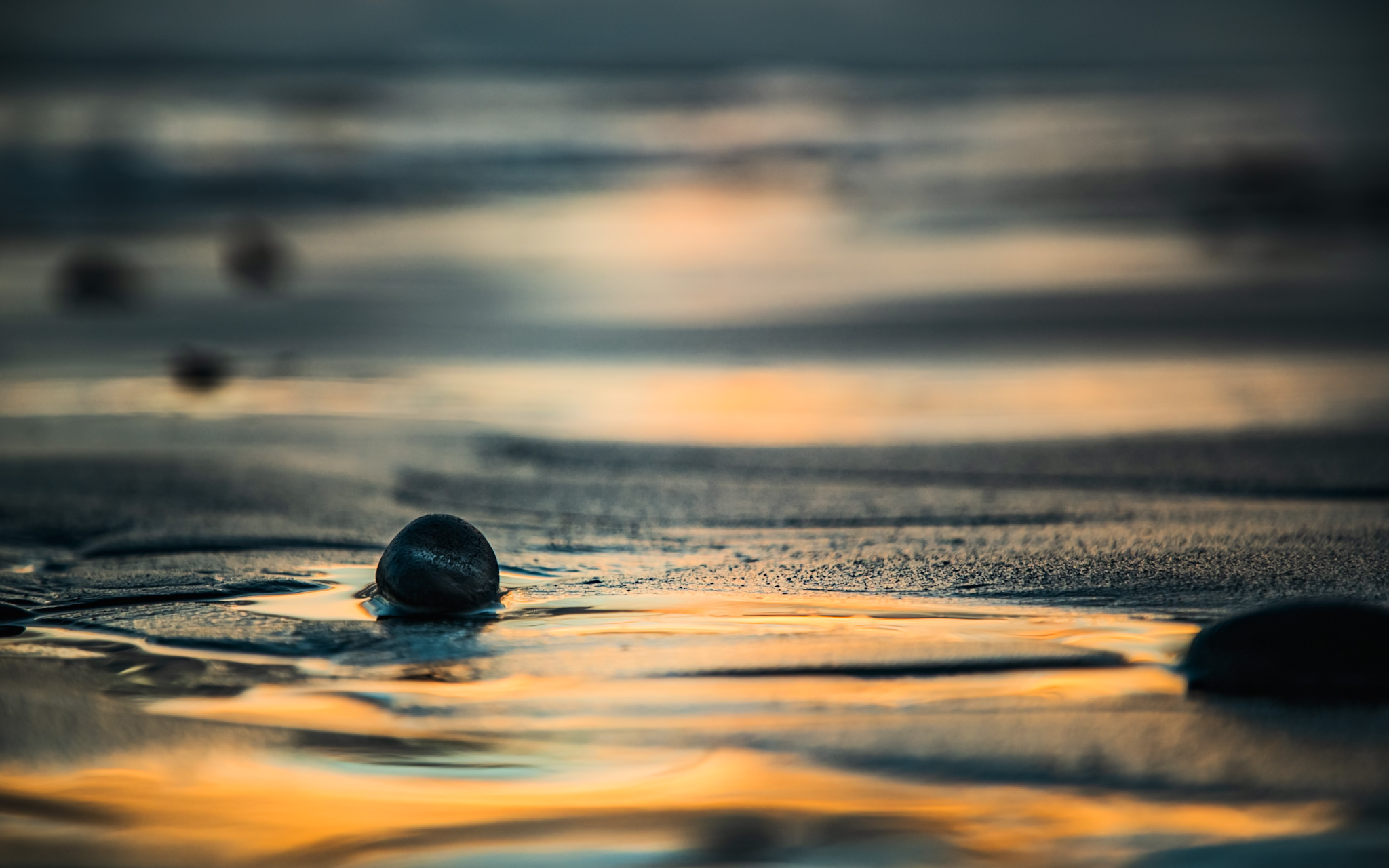 Pebbles on wet sand reflecting sunset light with blurred background of water and sky.