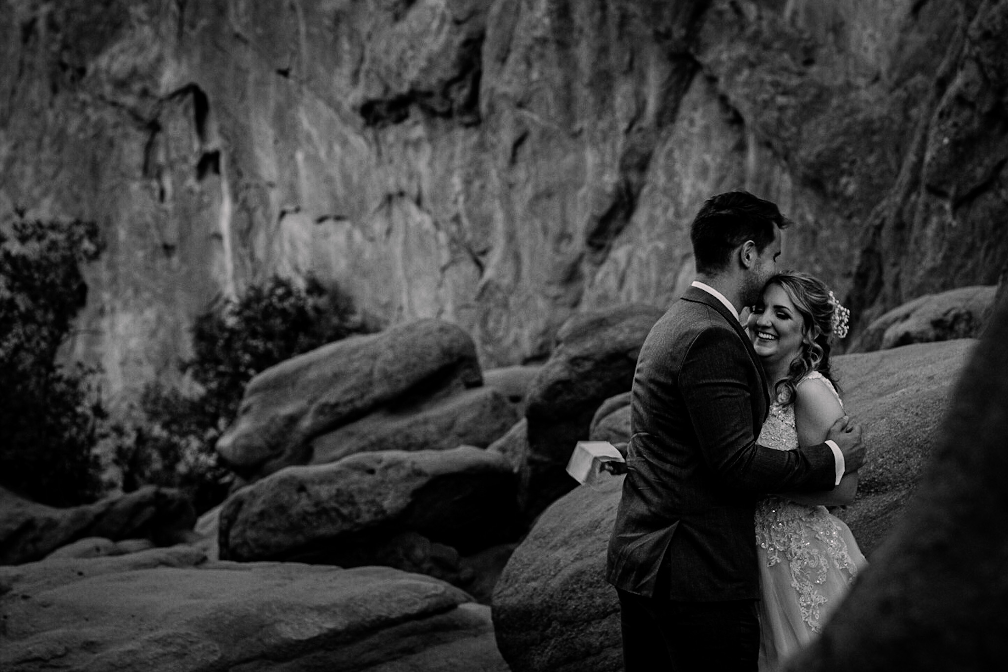 A couple in formal attire embracing outdoors with rocky terrain and cliffs in the background.
