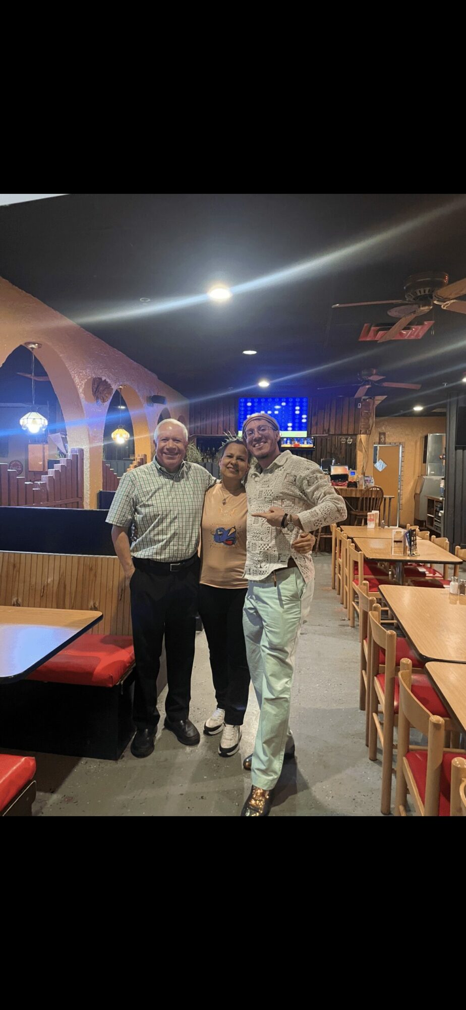 Three men standing together in a restaurant with tables and chairs, illuminated by colorful lighting and a bright ceiling light.
