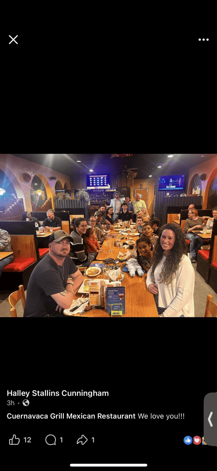 Group of people sitting at a long table in a restaurant with colorful decor and screens on walls.
