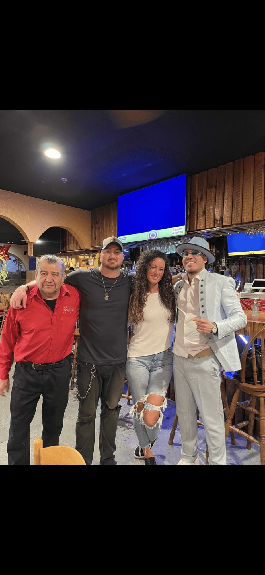 Four people standing together inside a restaurant, smiling, with wooden walls and television screens in the background.