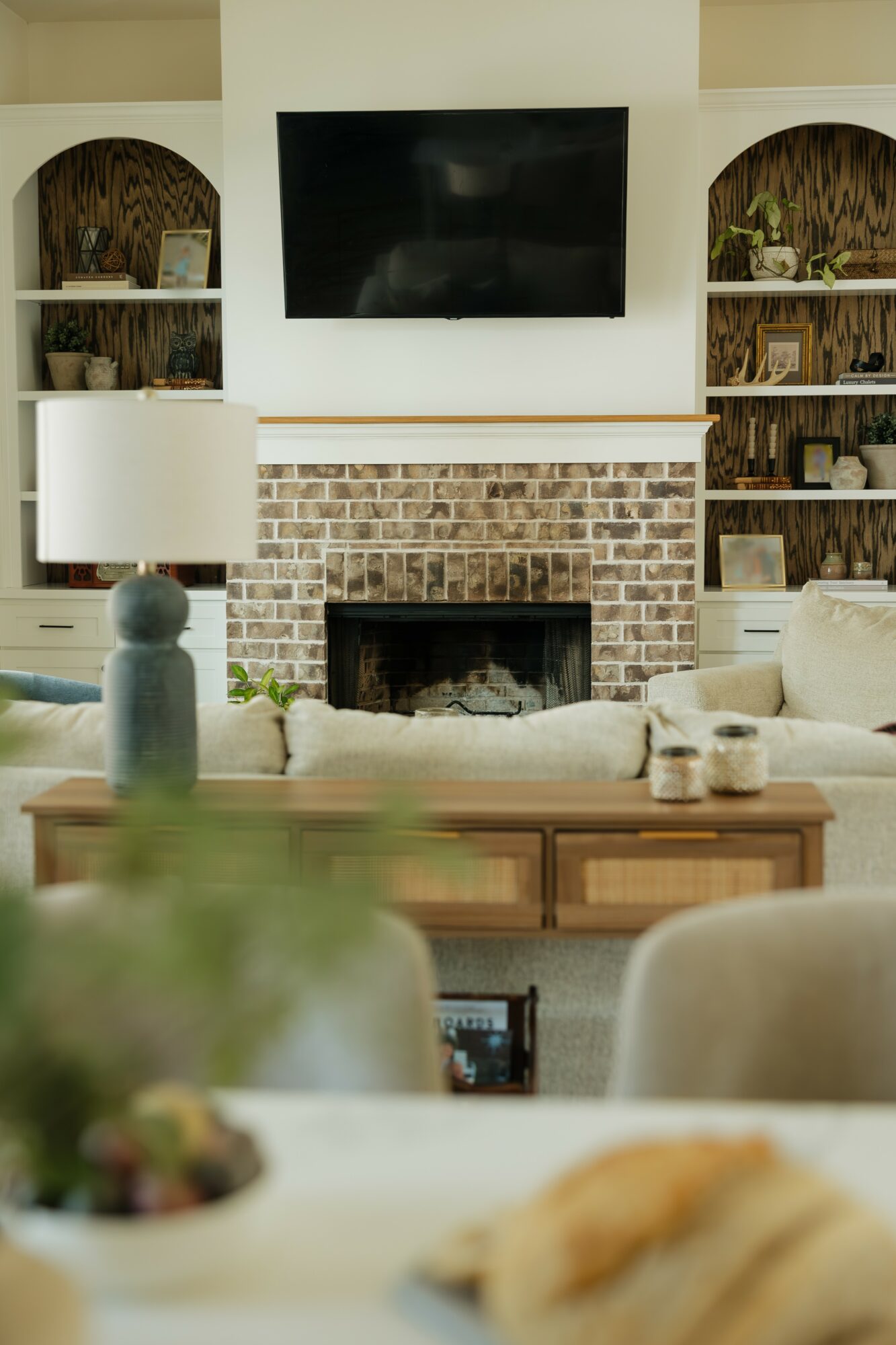 Living room with brick fireplace, wall-mounted TV, built-in shelves, and beige furniture.