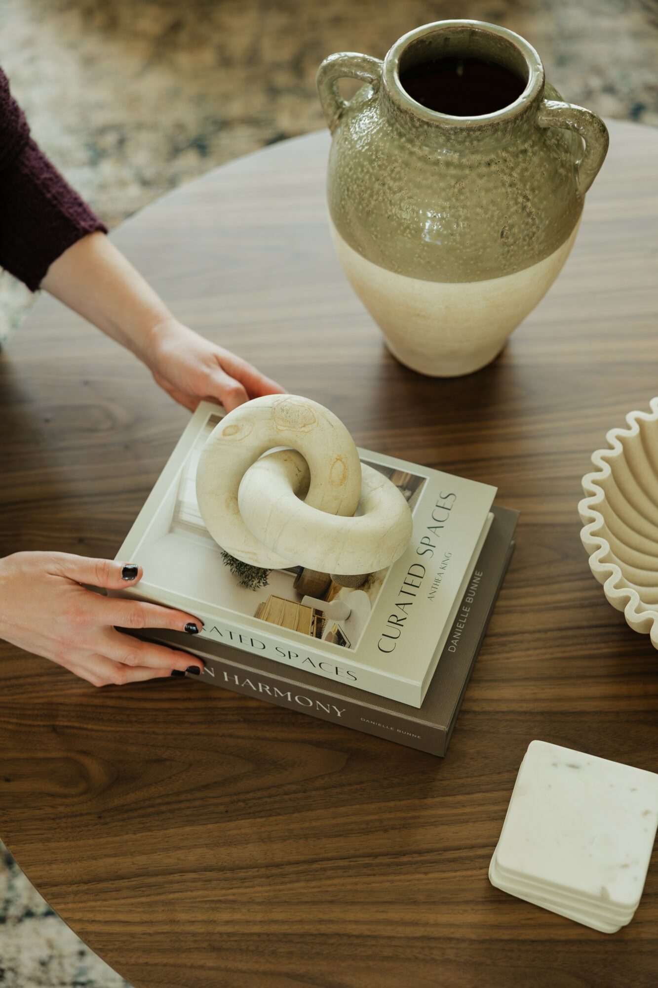 Person's hand holding a ceramic ring-shaped object over a stack of books on a wooden table, with a large ceramic vase nearby.
