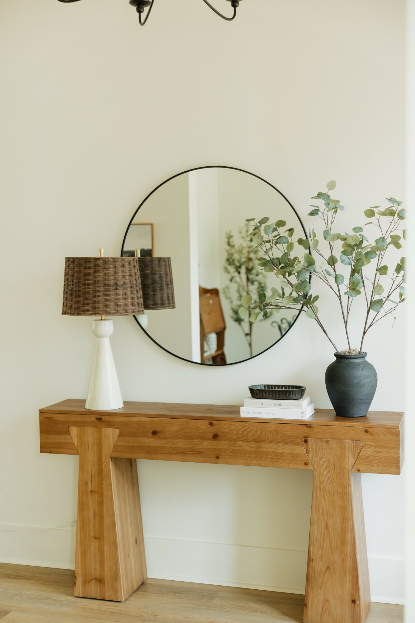 Wooden console table with a round mirror, lamp, books, and a vase with branches inside a room.
