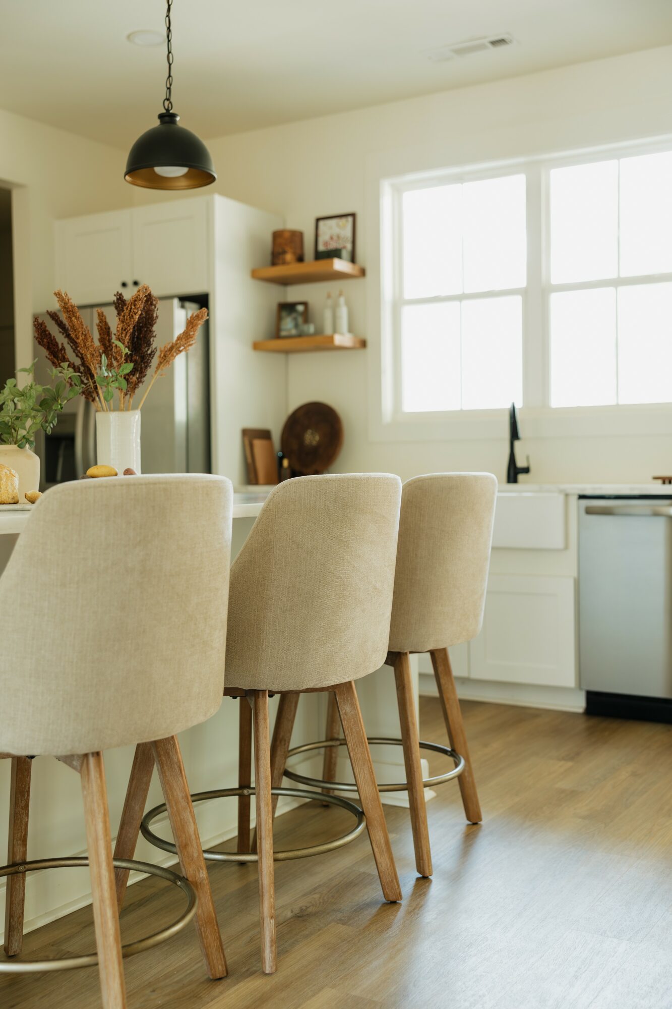 Kitchen with beige chairs around a wooden island, large window, and shelves with decor items.