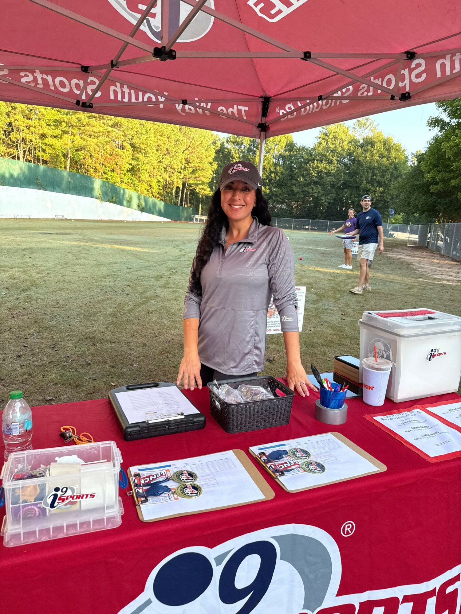 Woman standing behind a table under a red canopy outdoors, with people and trees in background.