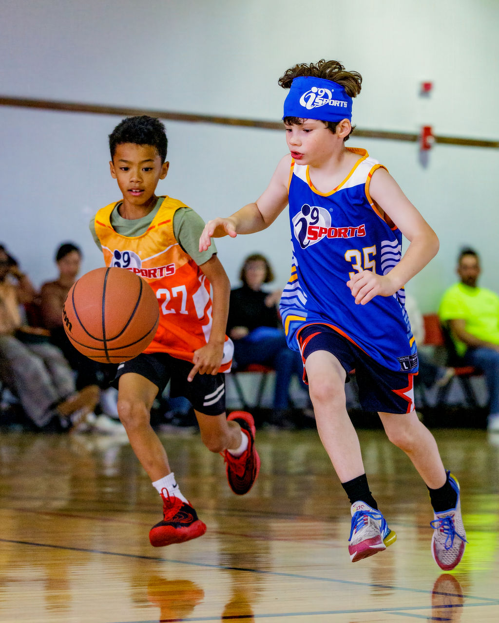 Two young boys playing basketball indoors, one in an orange jersey and the other in a blue jersey, running on the court.