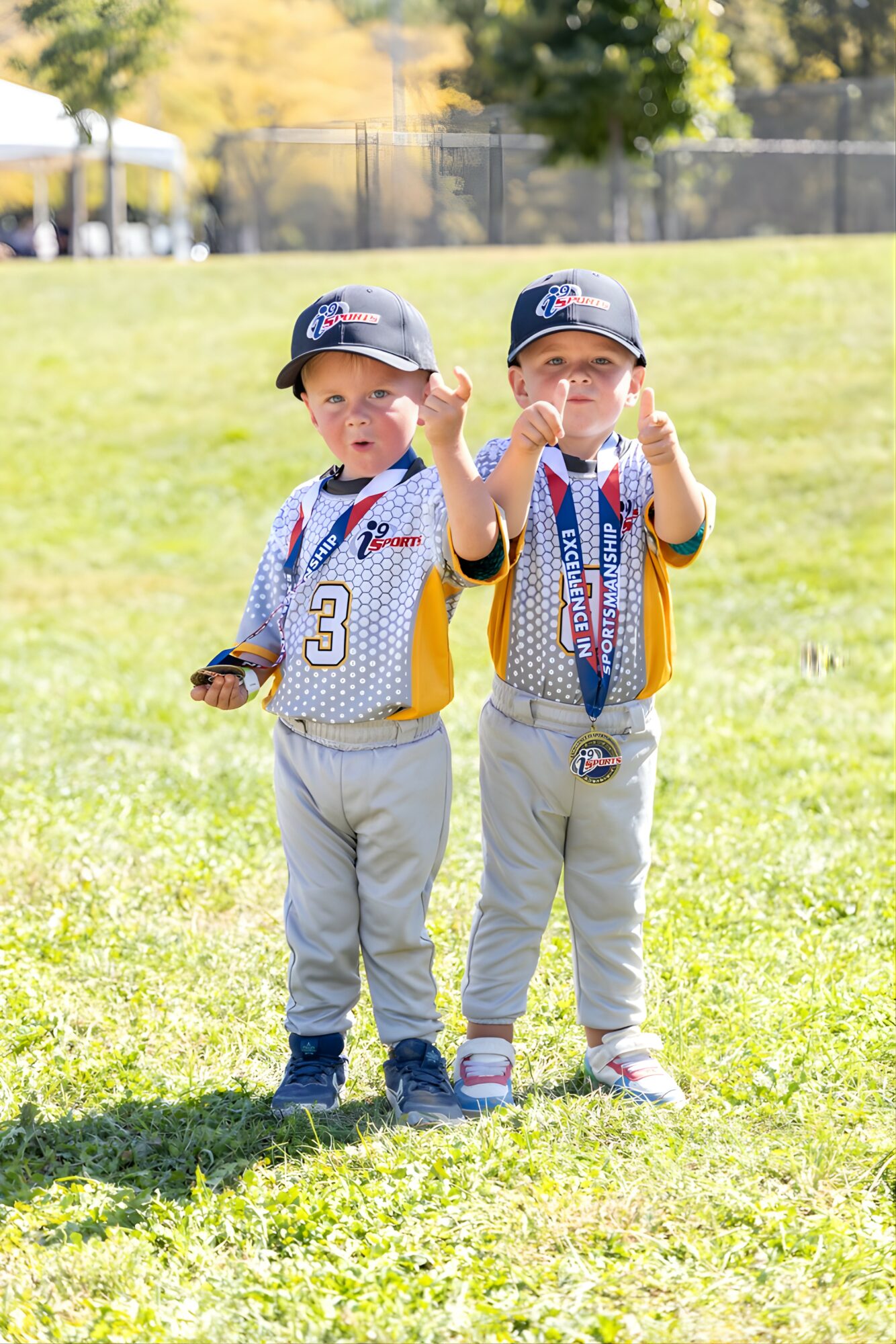 Two young boys in baseball uniforms standing on grass, one with a medal around his neck, outdoors on a sunny day.