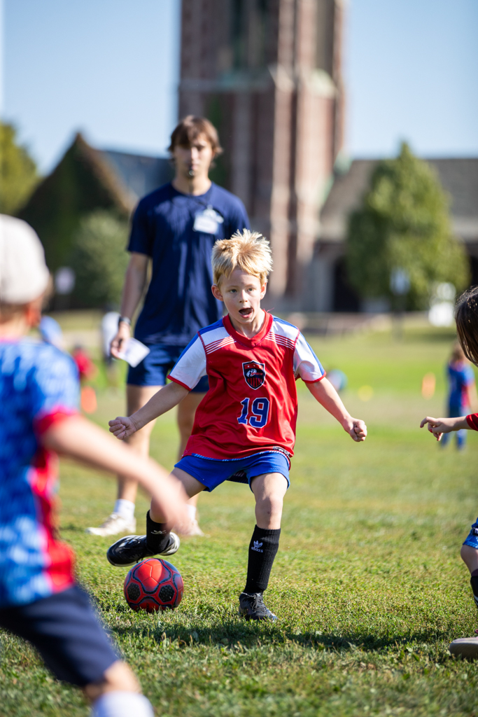 Child in red and blue soccer uniform kicking a ball on a grassy field, with a person and trees in background.