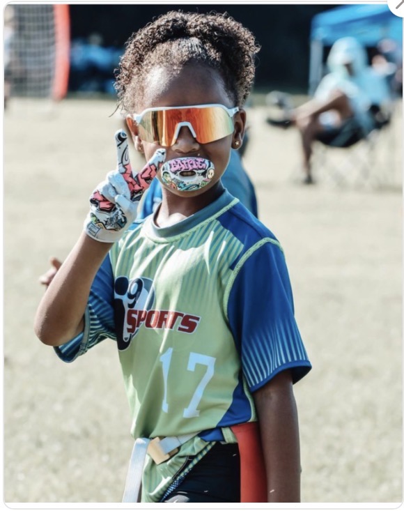 Child wearing sports uniform, sunglasses, and mouthguard, standing outdoors on grass field.