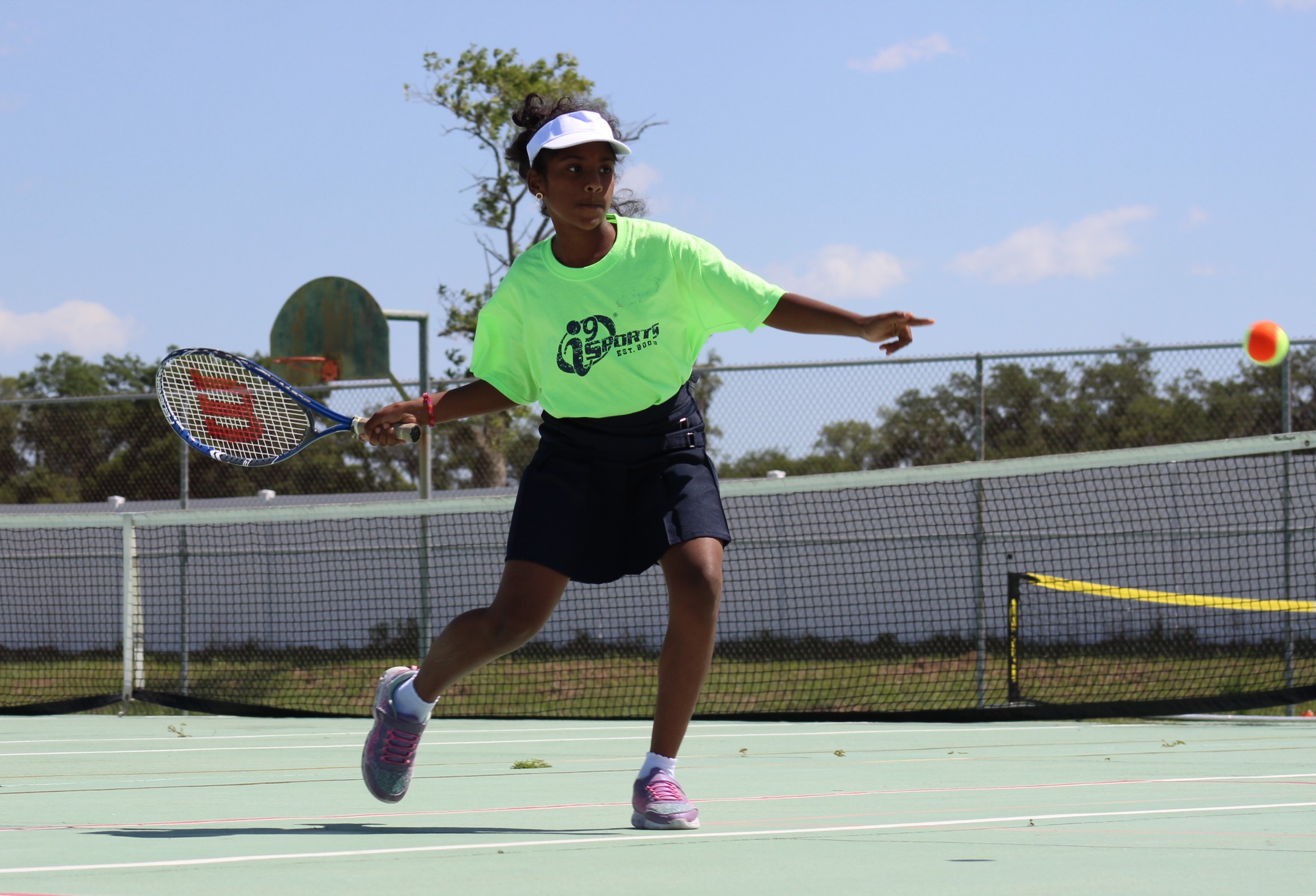 Child playing tennis on outdoor court, reaching to hit ball with racket, wearing bright green shirt and white cap.