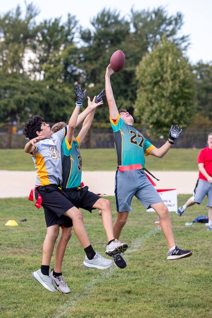 Three children playing football outdoors, jumping to catch or reach for the ball, with trees and a field in the background.