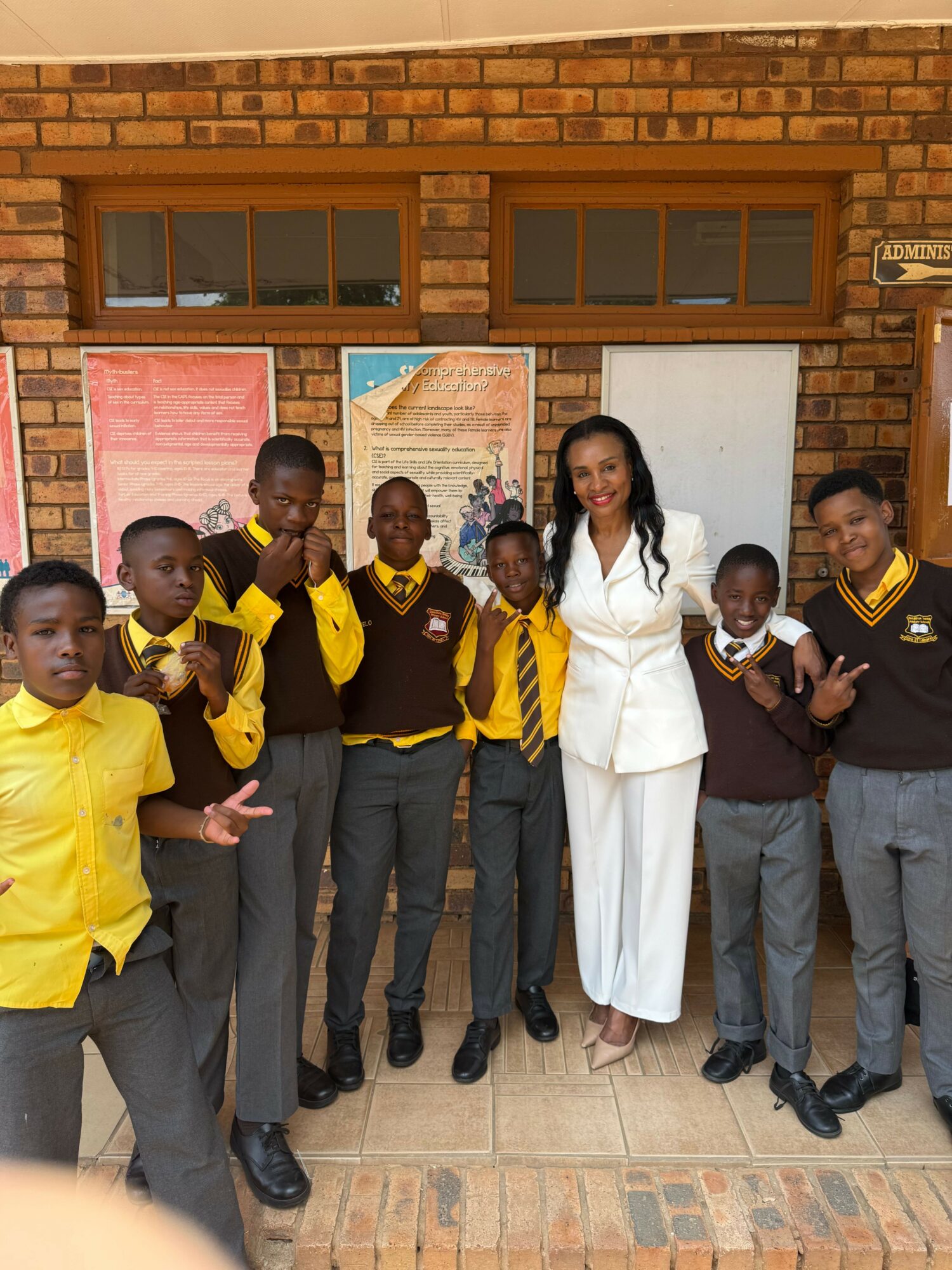 Group of children and a woman standing in front of a brick wall with posters and windows.