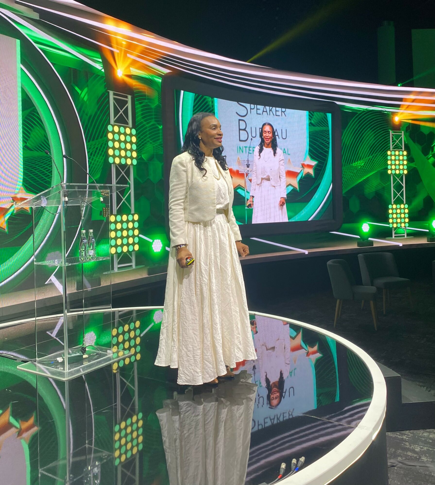 Woman in white dress standing on stage with large screen behind her displaying her image, stage lit with green and yellow lights.