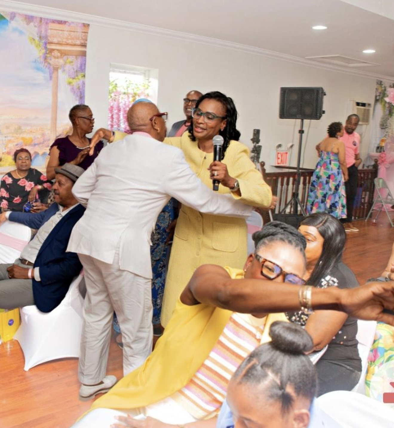 People dancing and socializing indoors, woman in yellow dress holding microphone, others seated and standing, colorful decorations.