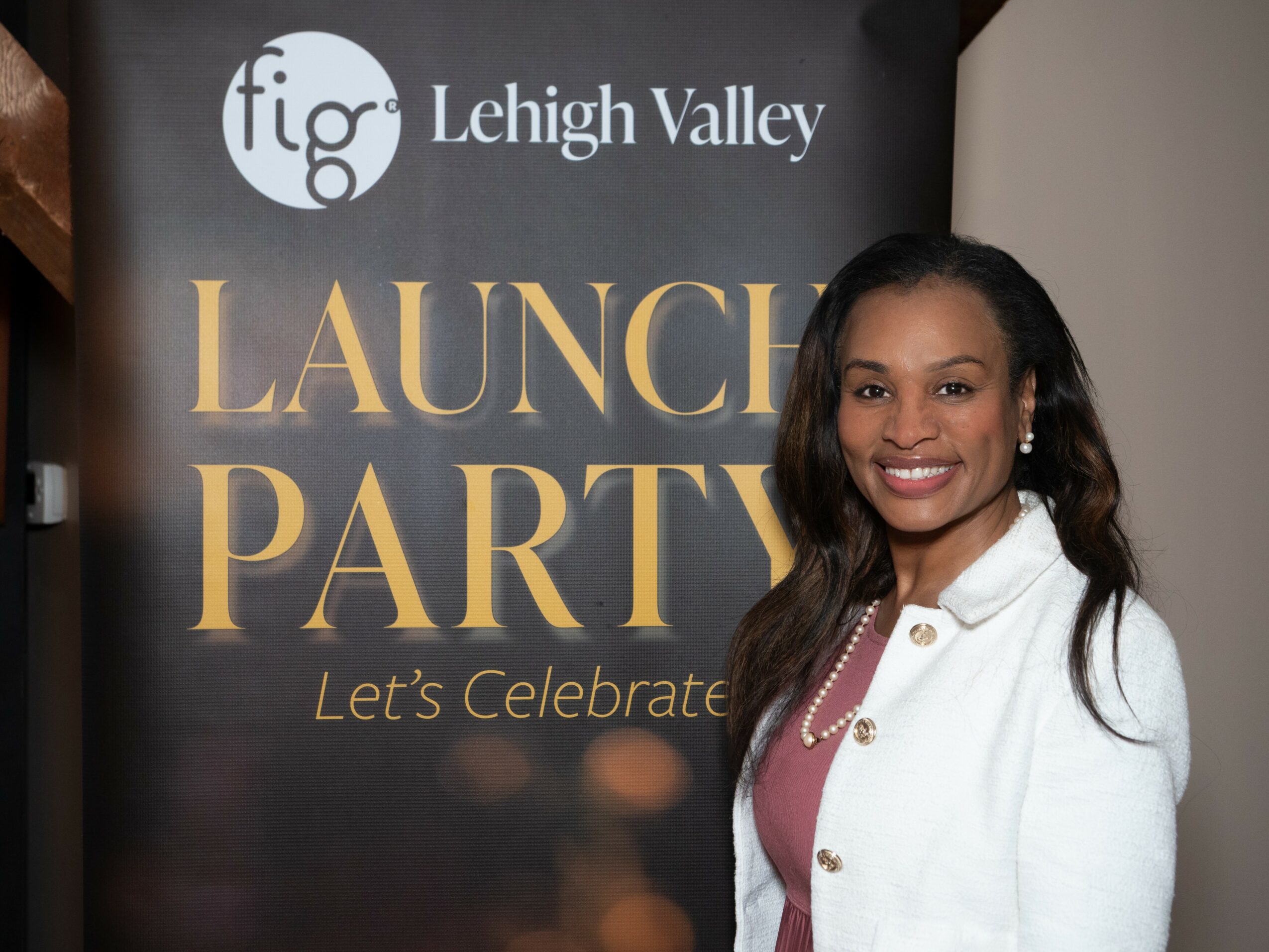 Smiling woman in white jacket standing in front of a black banner with gold text at an event.