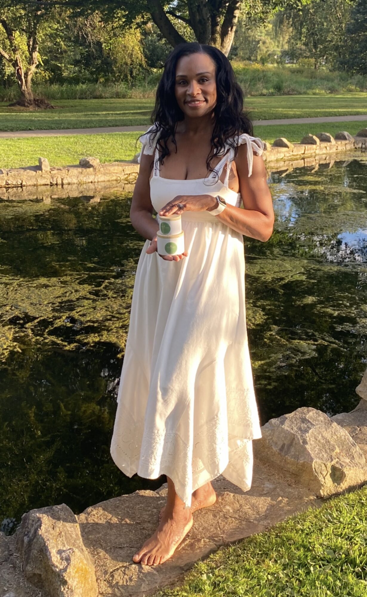 Woman in white dress holding a drink standing on rocks near water in park.
