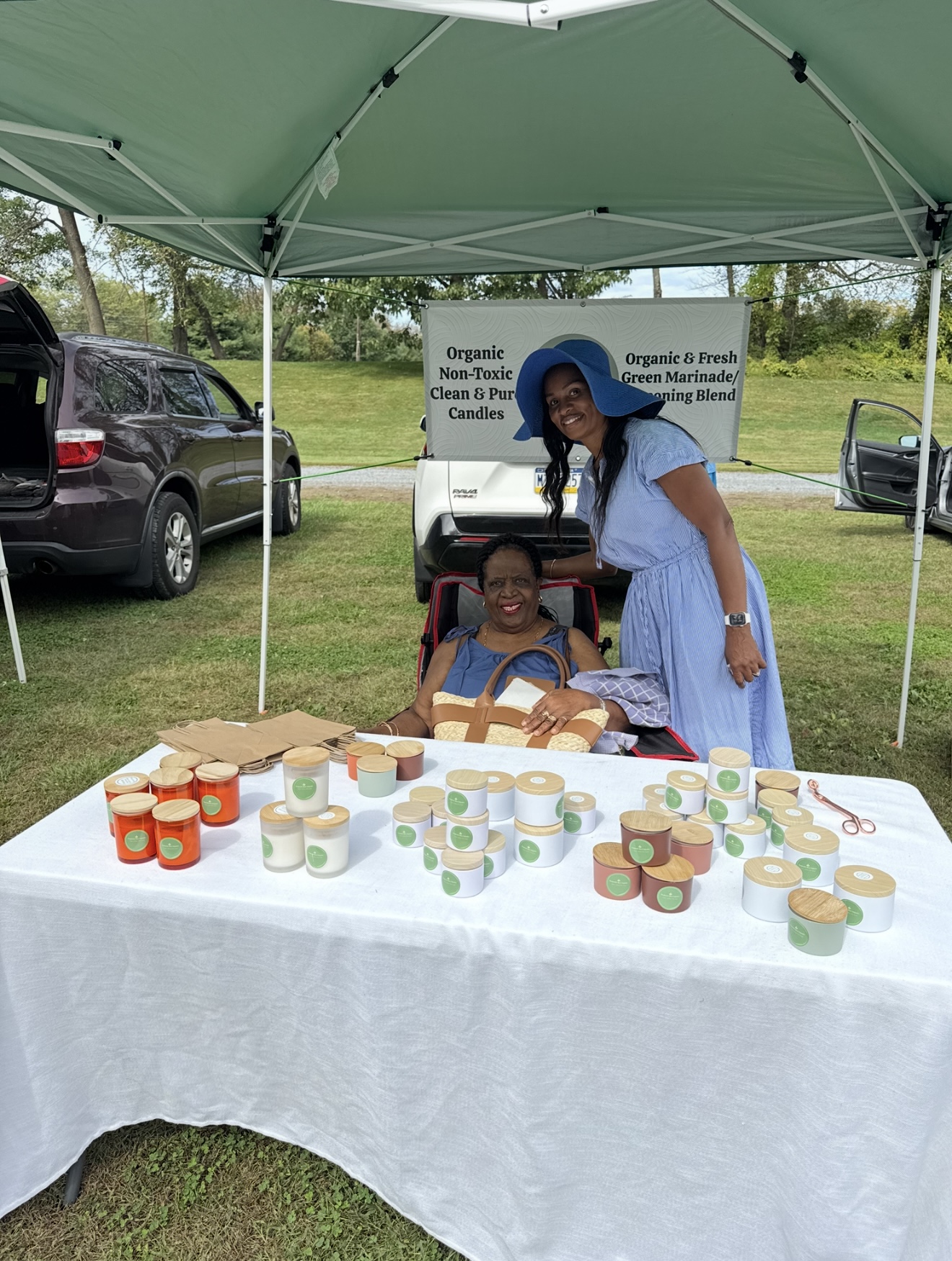 Two women at a table with jars of products under a green canopy outdoors, cars parked nearby, grassy area in background.