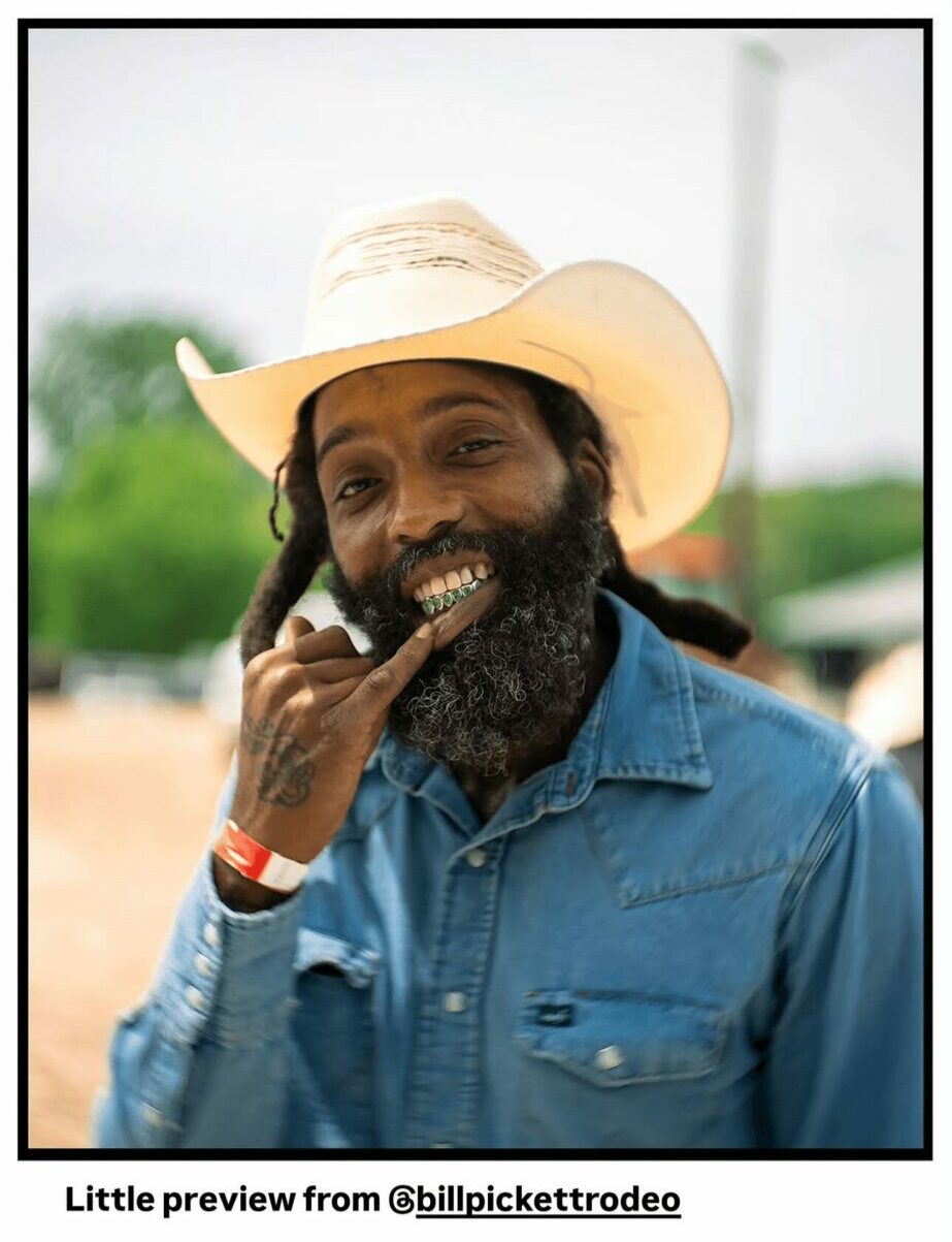 Man with a beard wearing a wide-brimmed hat and denim shirt, smiling outdoors.