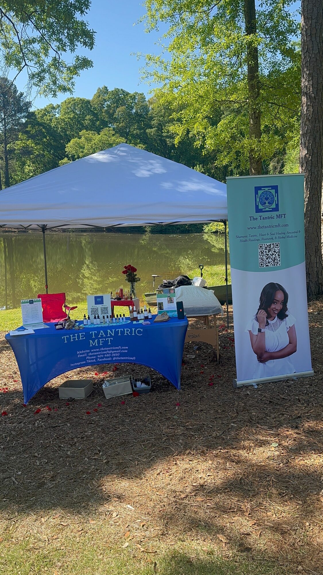 Outdoor booth with table covered in blue cloth, a white tent, and a vertical sign near a pond and trees.