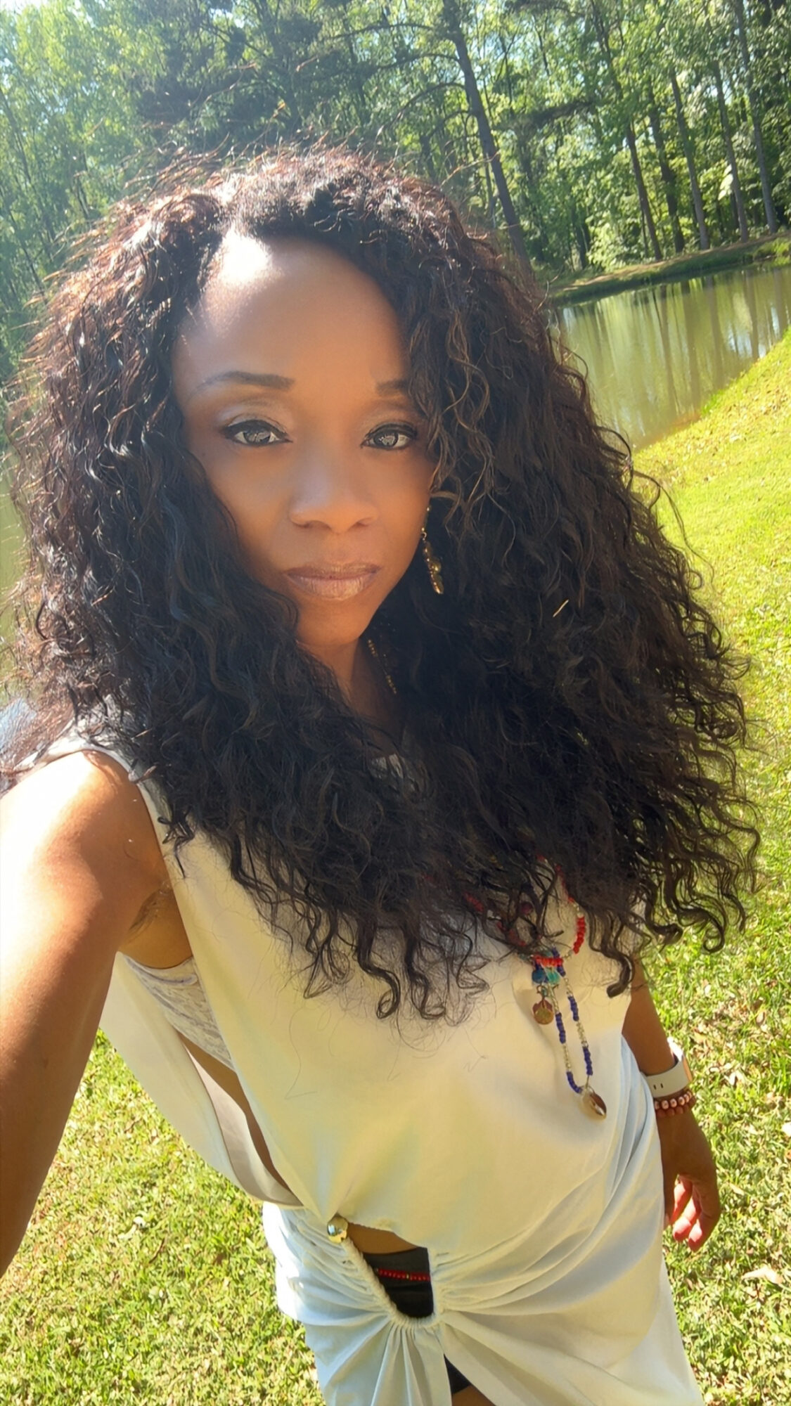 Woman with curly hair standing outdoors near a pond and trees, wearing a light-colored top.