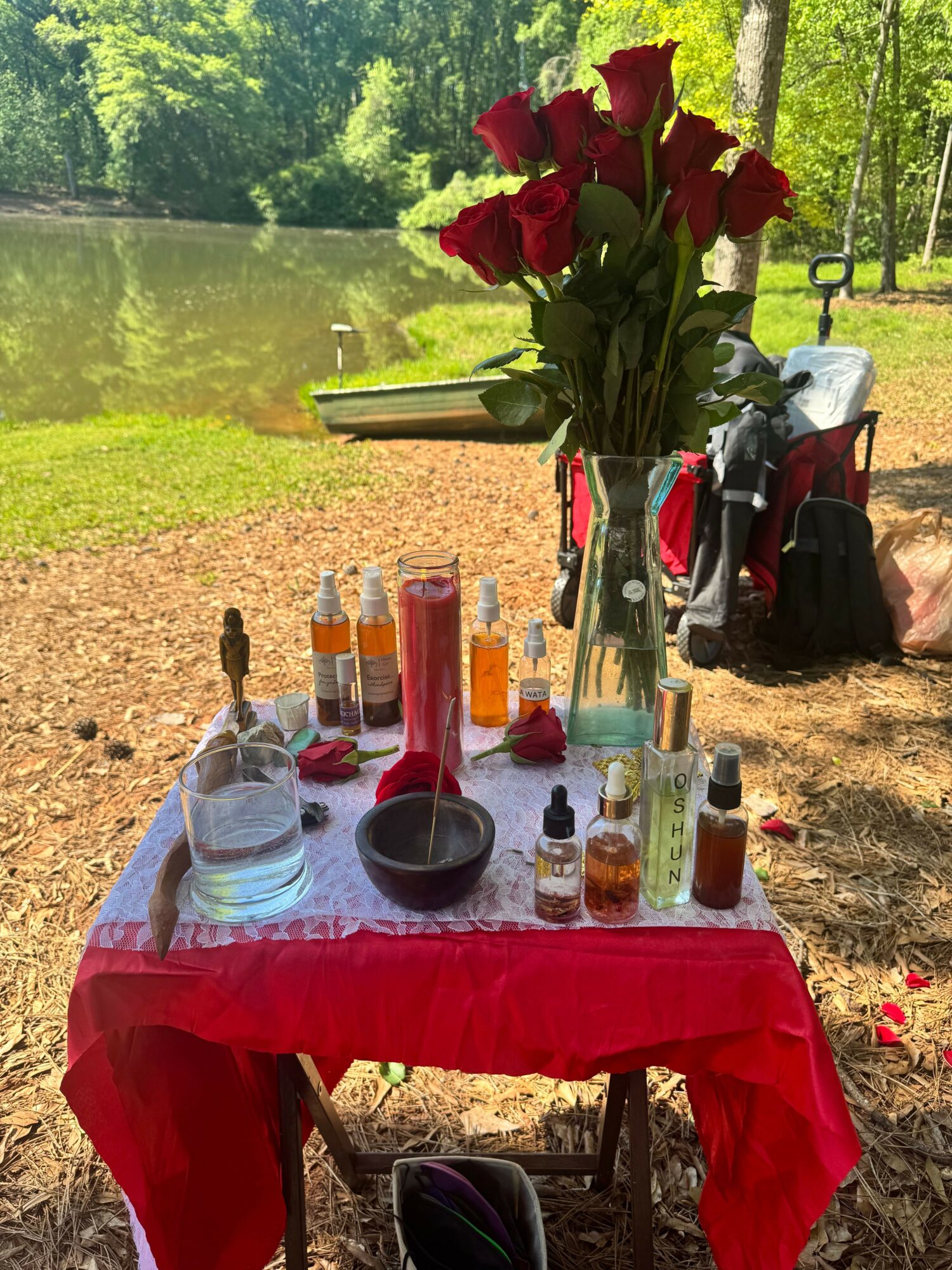 Table with red tablecloth, flowers, bottles, and bowls outdoors near a lake with trees.