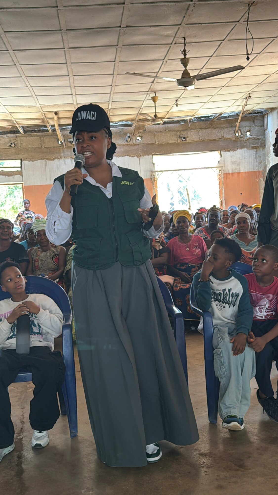 Girl in uniform speaking into microphone in a large indoor gathering with children seated behind her.