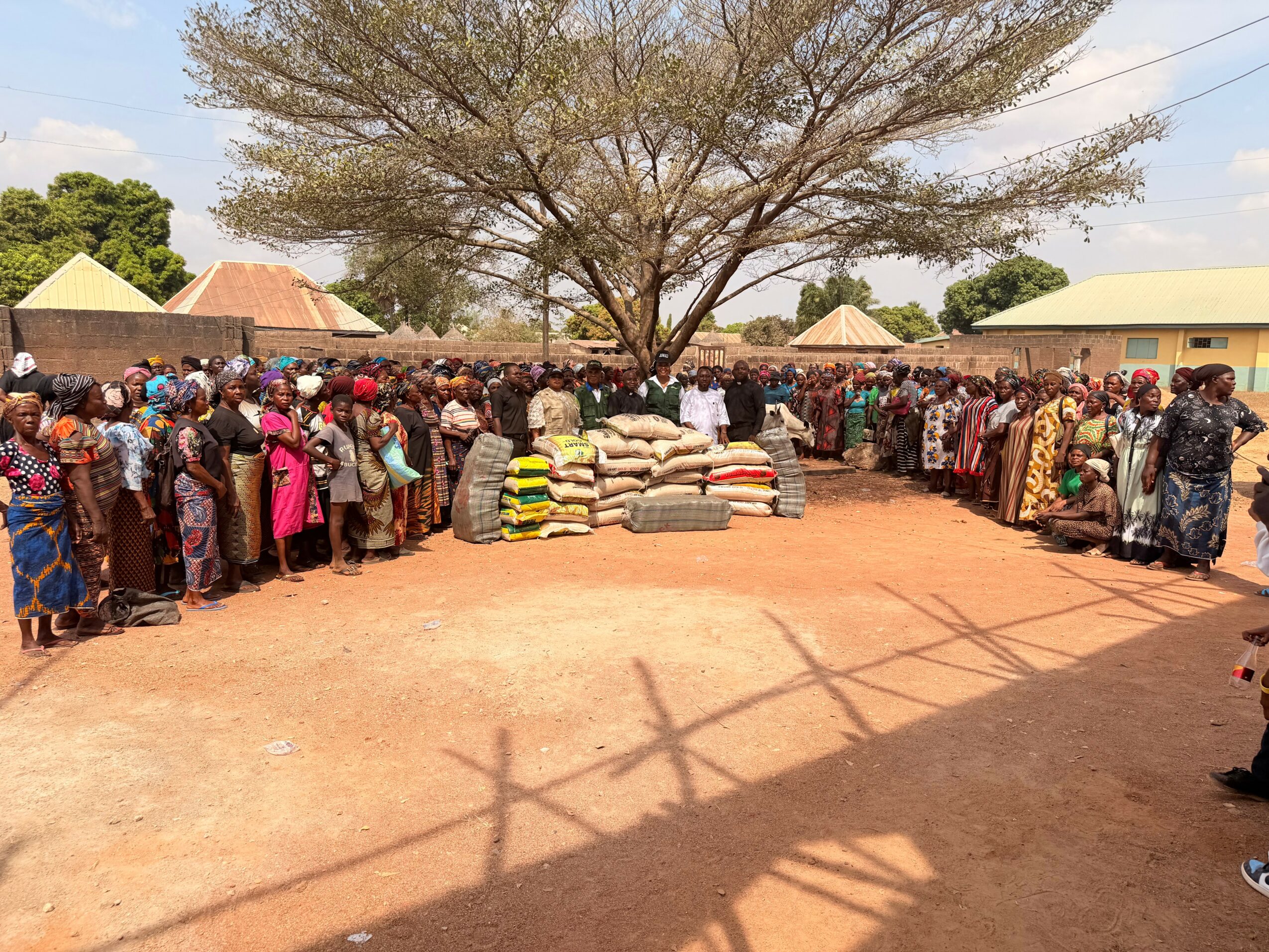 Large group of people gathered outdoors around stacks of bags and sacks, with a large tree in the background.