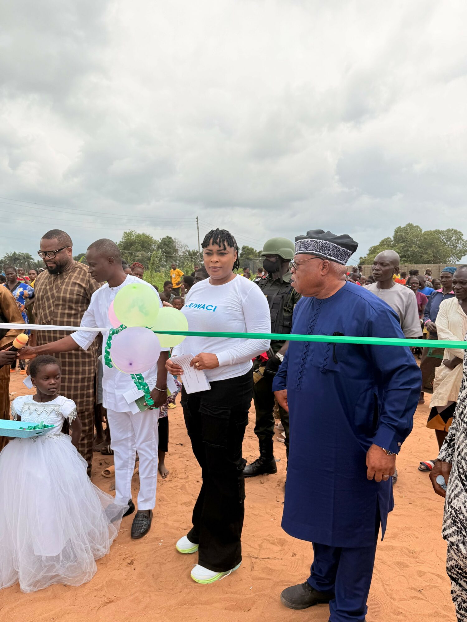 Group of people gathered outdoors for a ribbon-cutting event, with a young girl in a white dress and a man in traditional attire.