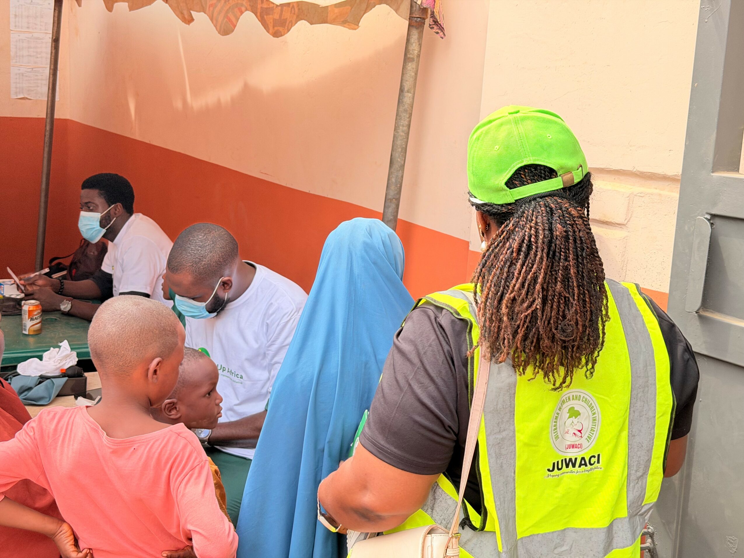 Person wearing a neon green helmet and yellow safety vest with a logo, facing a group of people sitting at a table.