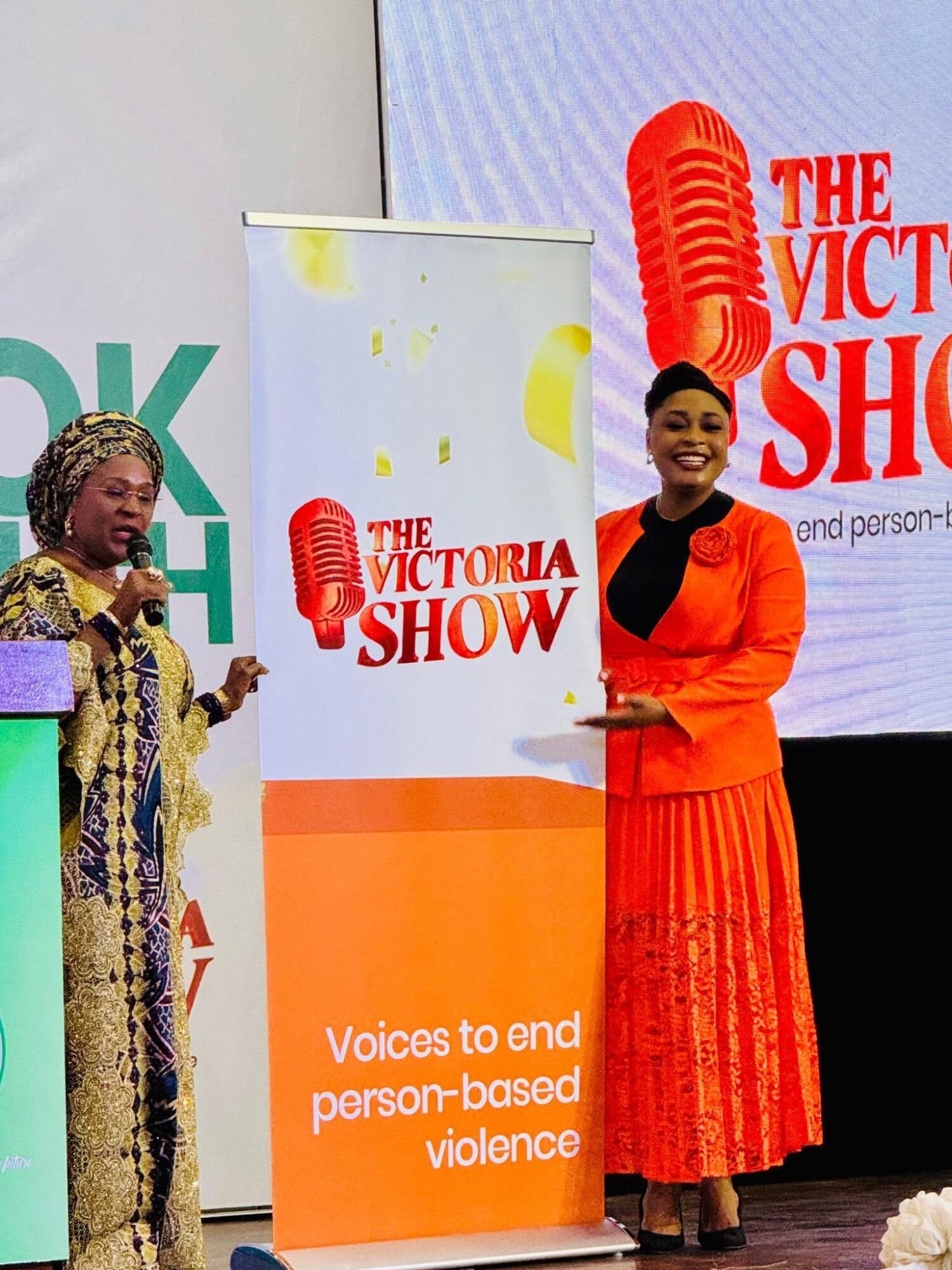 Two women standing on stage with a banner and screen behind them, promoting ending person-based violence.