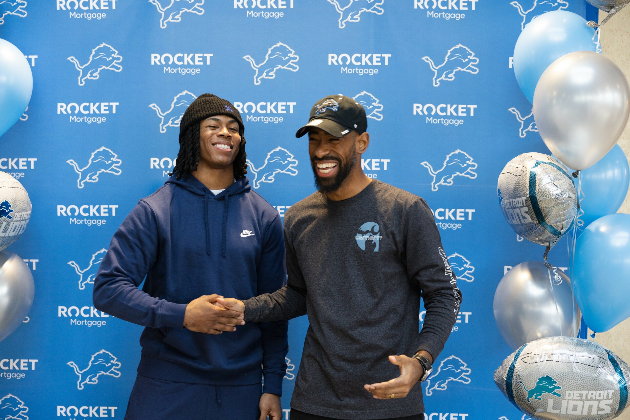 Two people shaking hands in front of a blue backdrop with Rocket Mortgage and Detroit Lions logos, smiling.