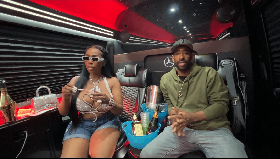 Two people sitting inside a luxury vehicle with black interior, drinks and snacks on the side, night sky visible through window.