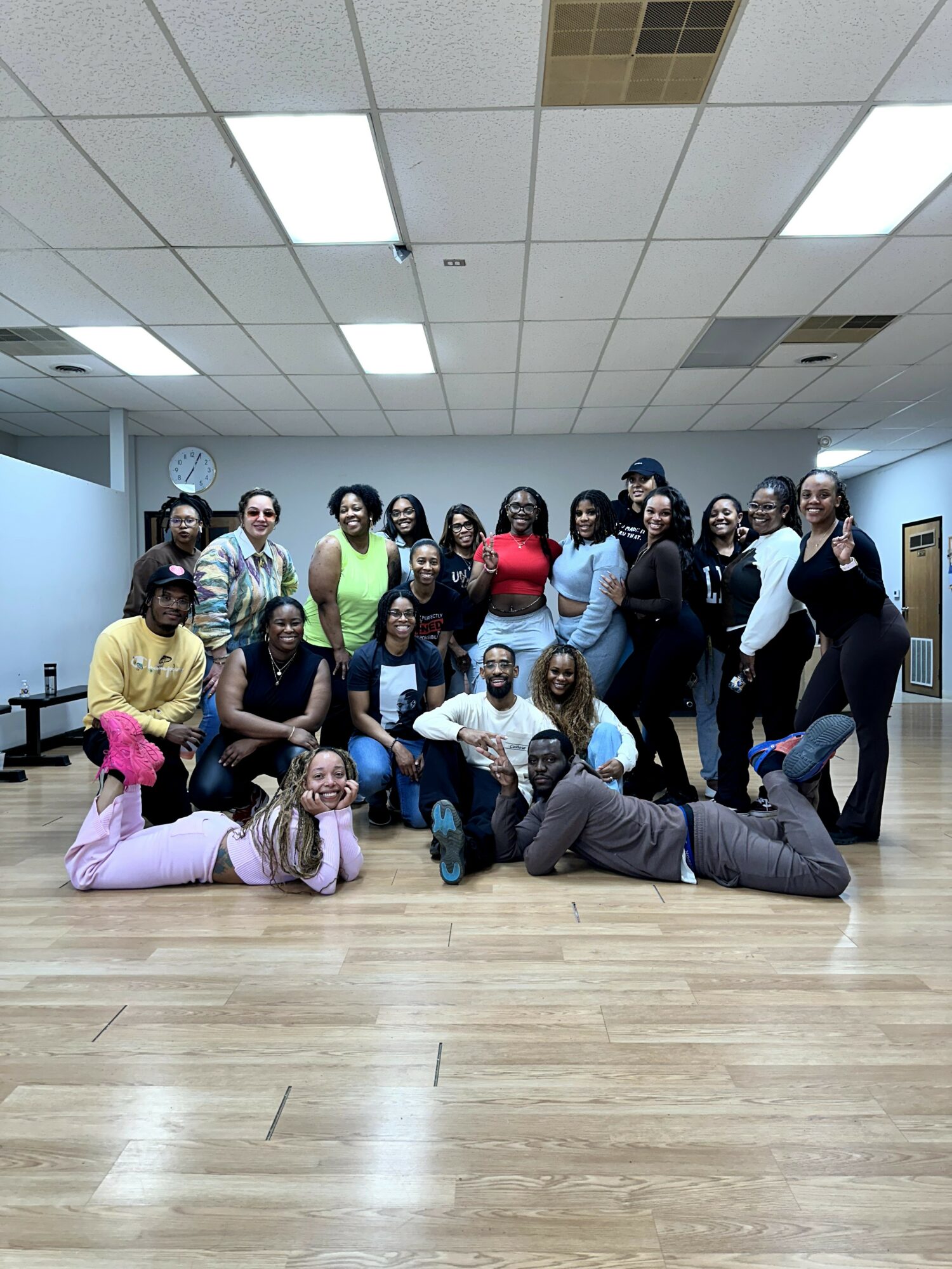Group of 17 people posing indoors, some standing and some lying on the floor, in a room with ceiling lights.