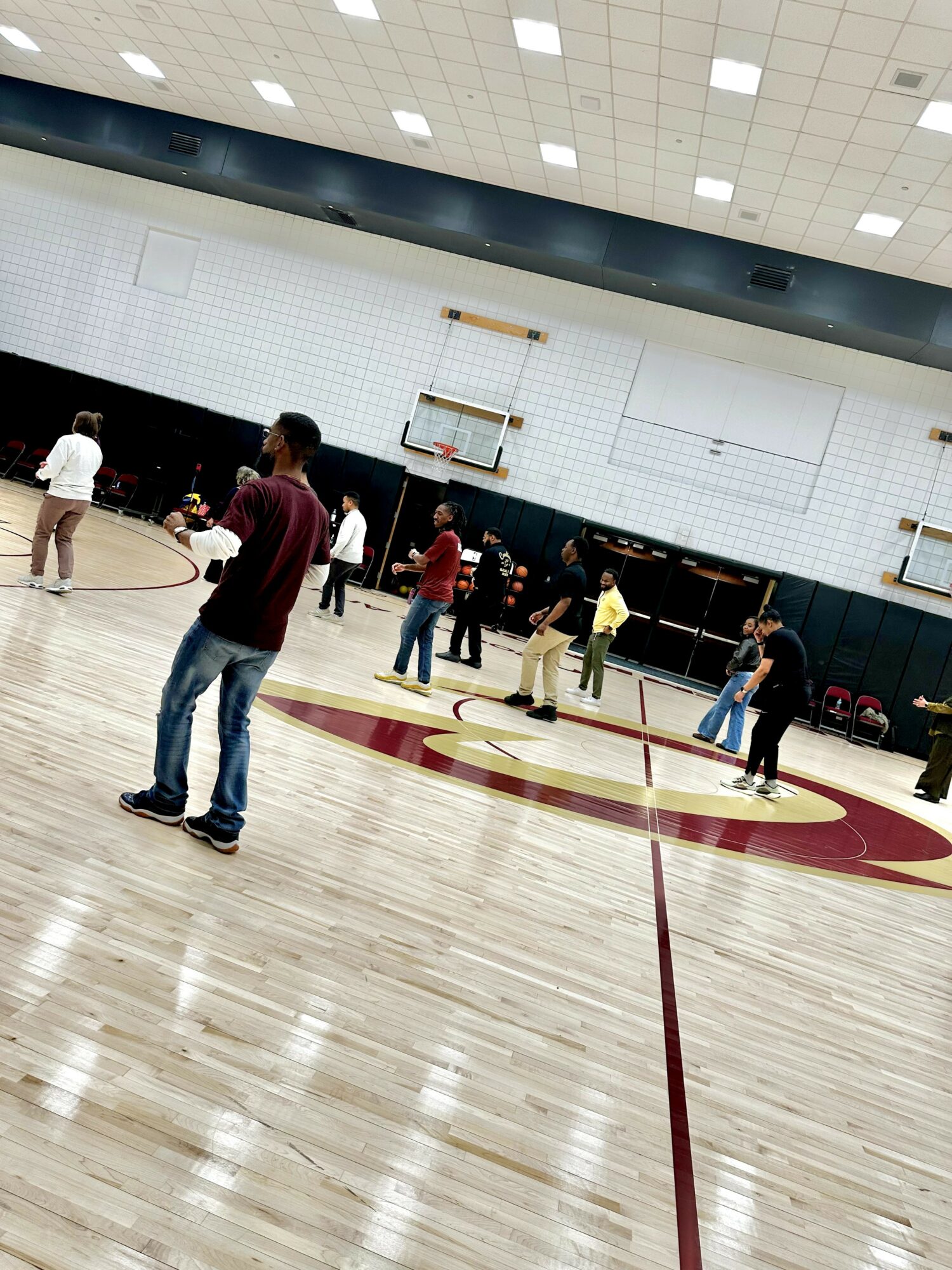 People playing basketball in an indoor gymnasium with a wooden floor and a basketball hoop.
