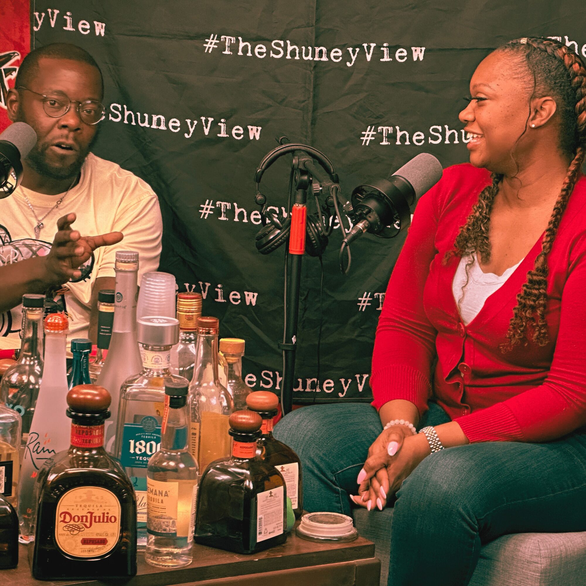 Two people sit at a table with various bottles, microphones, and a backdrop with text, engaged in conversation.