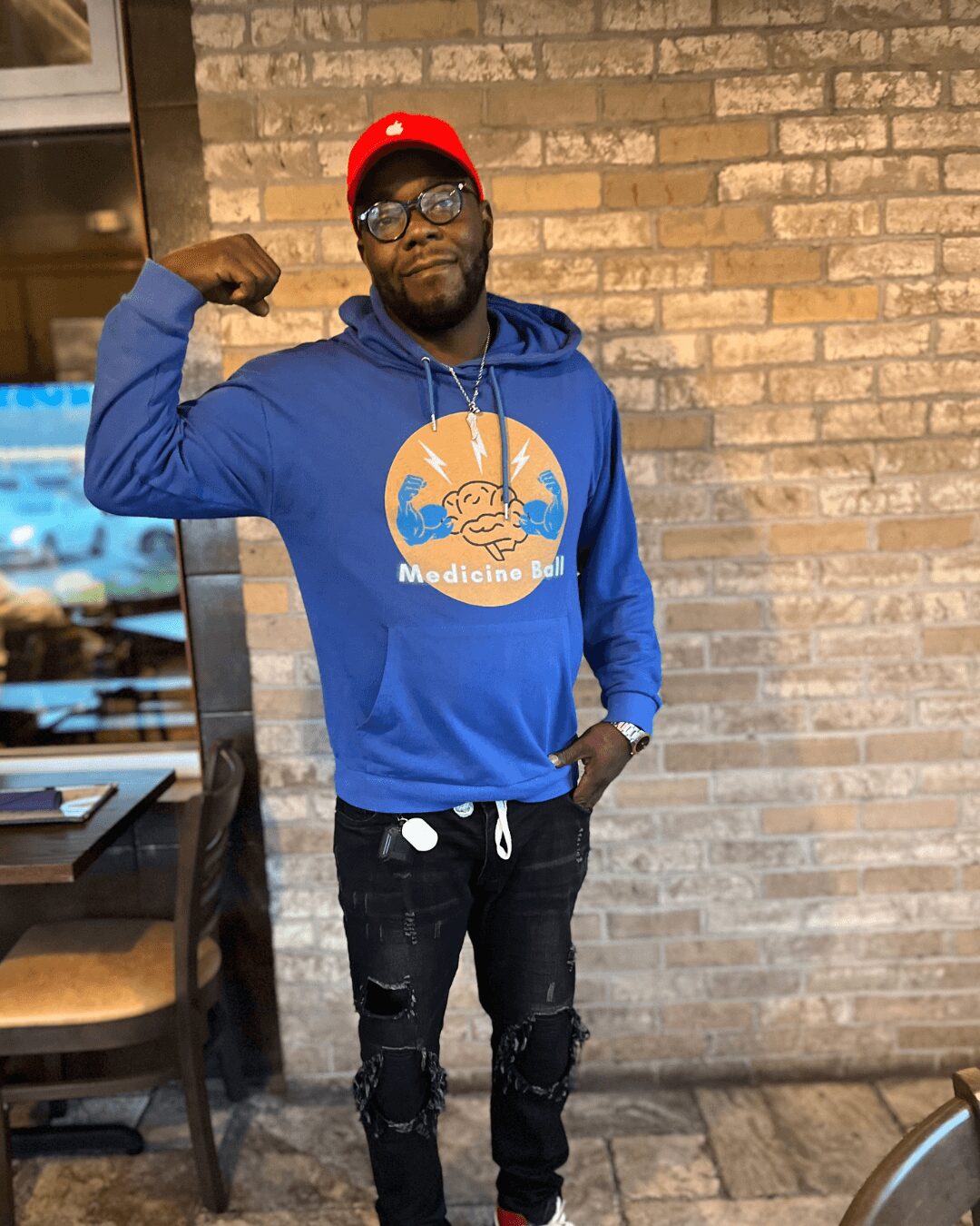 Man wearing glasses, red cap, blue hoodie with logo, black ripped jeans, standing against brick wall, flexing arm.