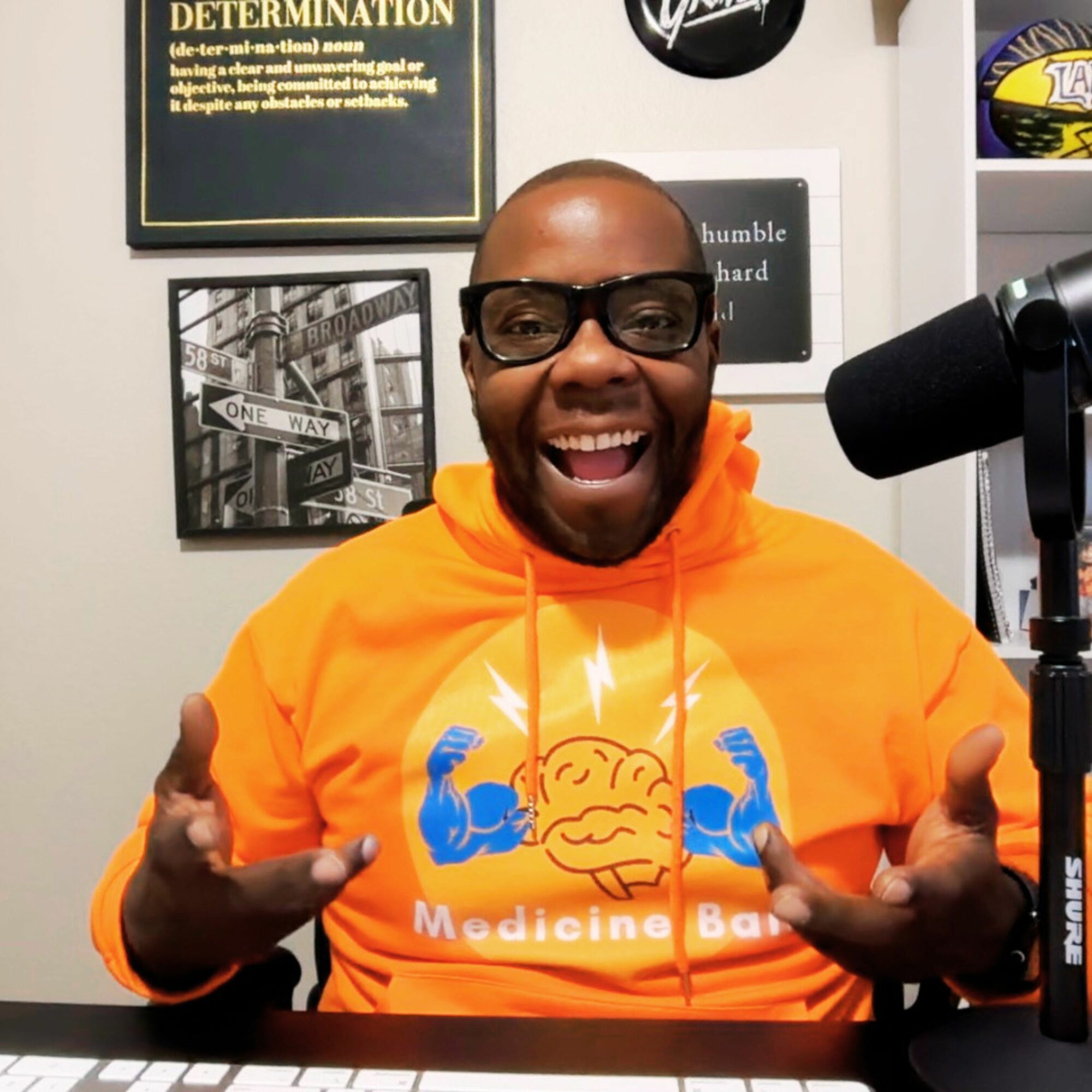Person smiling, wearing glasses and an orange hoodie with a cloud and muscle arms graphic, in a room with framed pictures and a microphone.