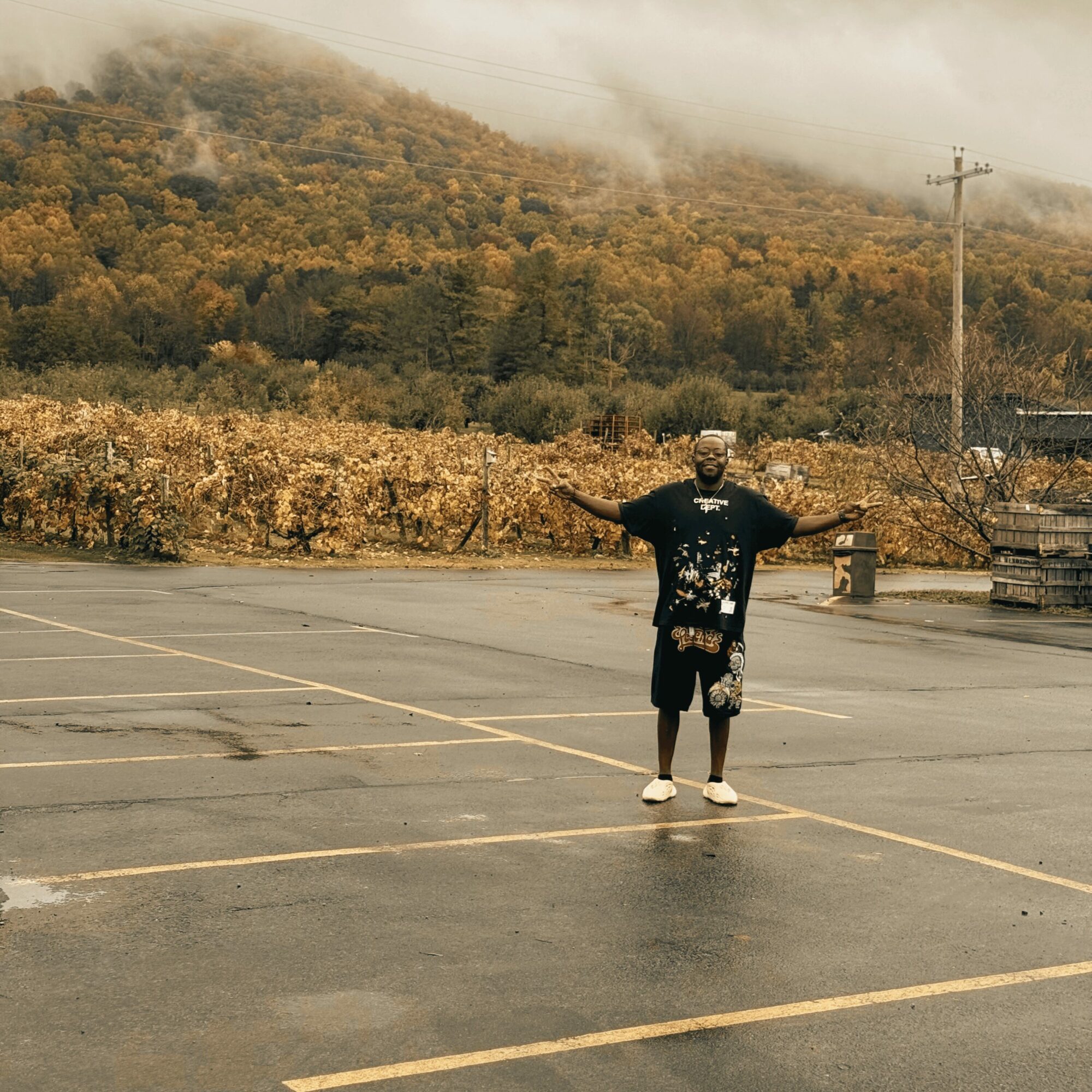Person standing in an empty parking lot with arms outstretched, autumn landscape with trees and hills in background.