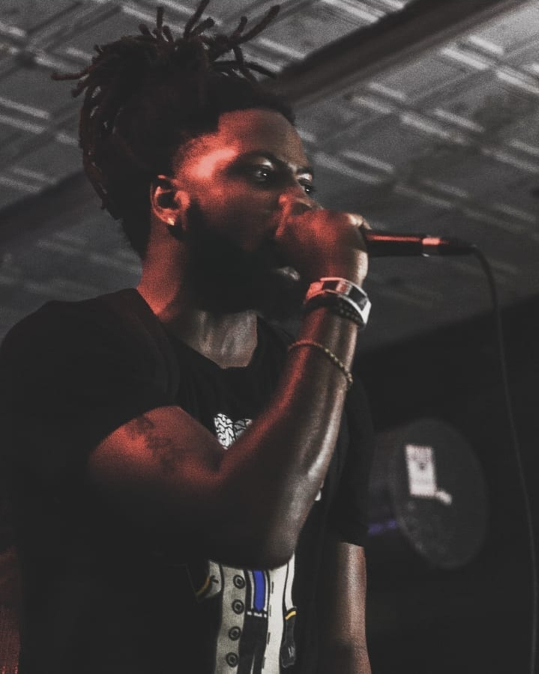 Person with dreadlocks singing into a microphone on stage, dark background, focused expression.
