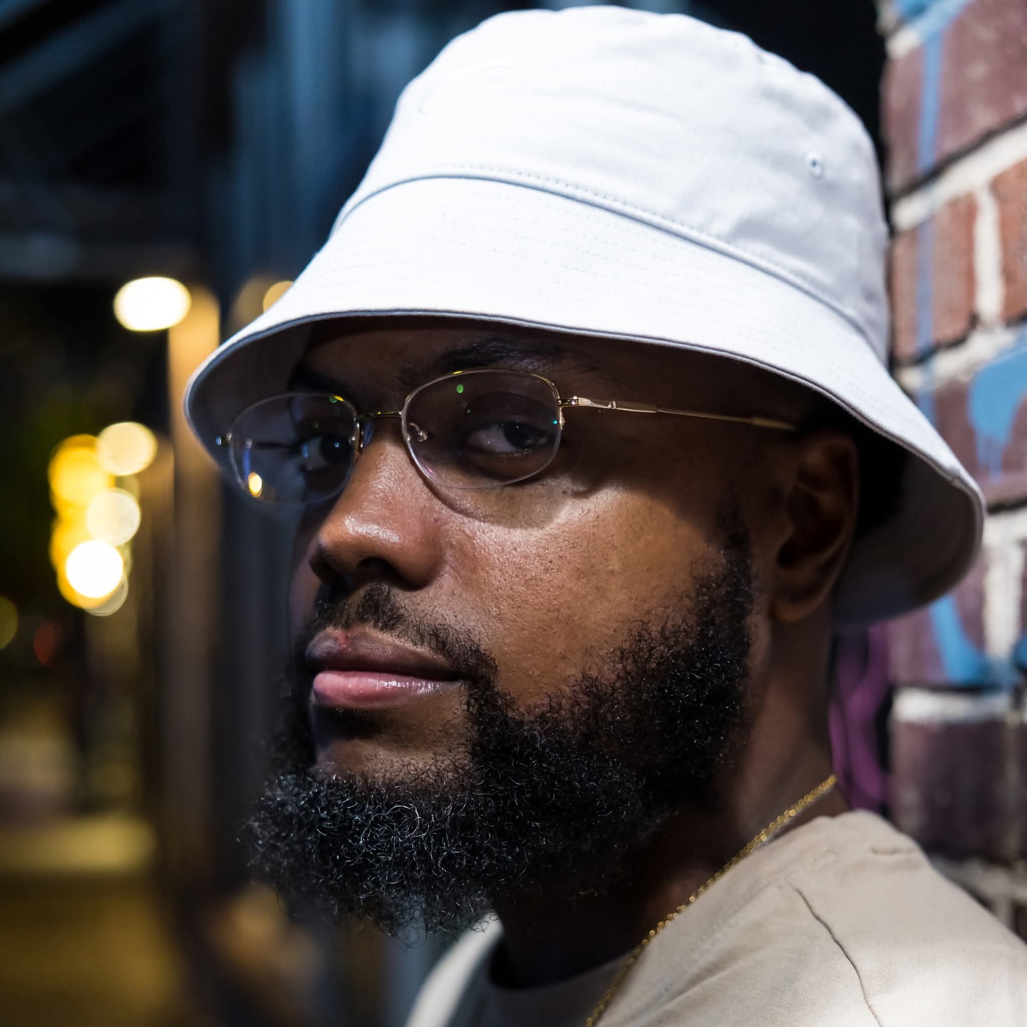 Man wearing white bucket hat and glasses, standing against a brick wall at night.