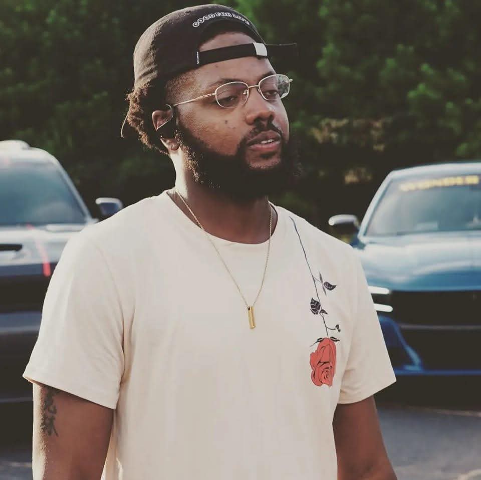 Man with glasses, beard, and cap standing outdoors near parked cars.