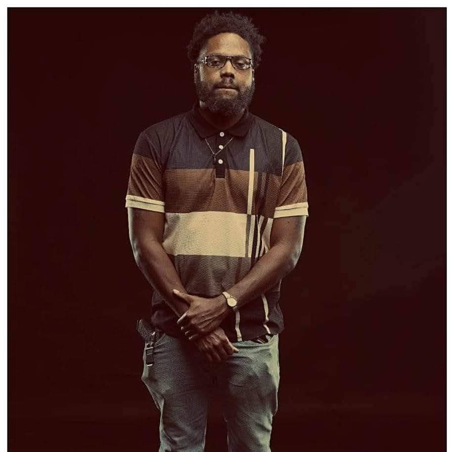 Man with glasses and beard wearing a striped polo shirt and jeans, standing against a dark background.