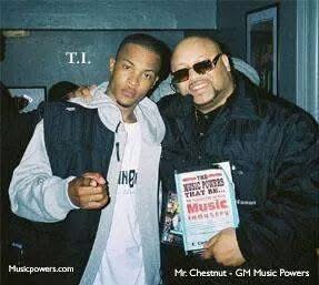 Two men standing together, one holding a booklet, in a room with blue walls and framed pictures.