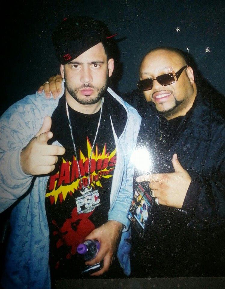 Two men posing together, one pointing at the camera, the other smiling with sunglasses, dark background with small stars.