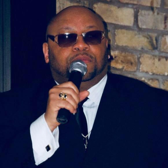 Man wearing sunglasses, holding microphone, dressed in dark suit and white shirt, standing against brick wall.